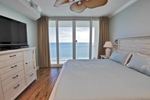 A hotel room with a large bed, a TV on a dresser, a nightstand with a lamp, and a balcony with a view of the sea.