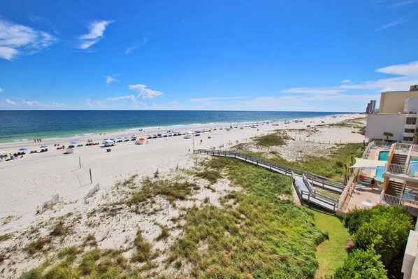 A beach with a wooden walkway leading to a building with a pool.
