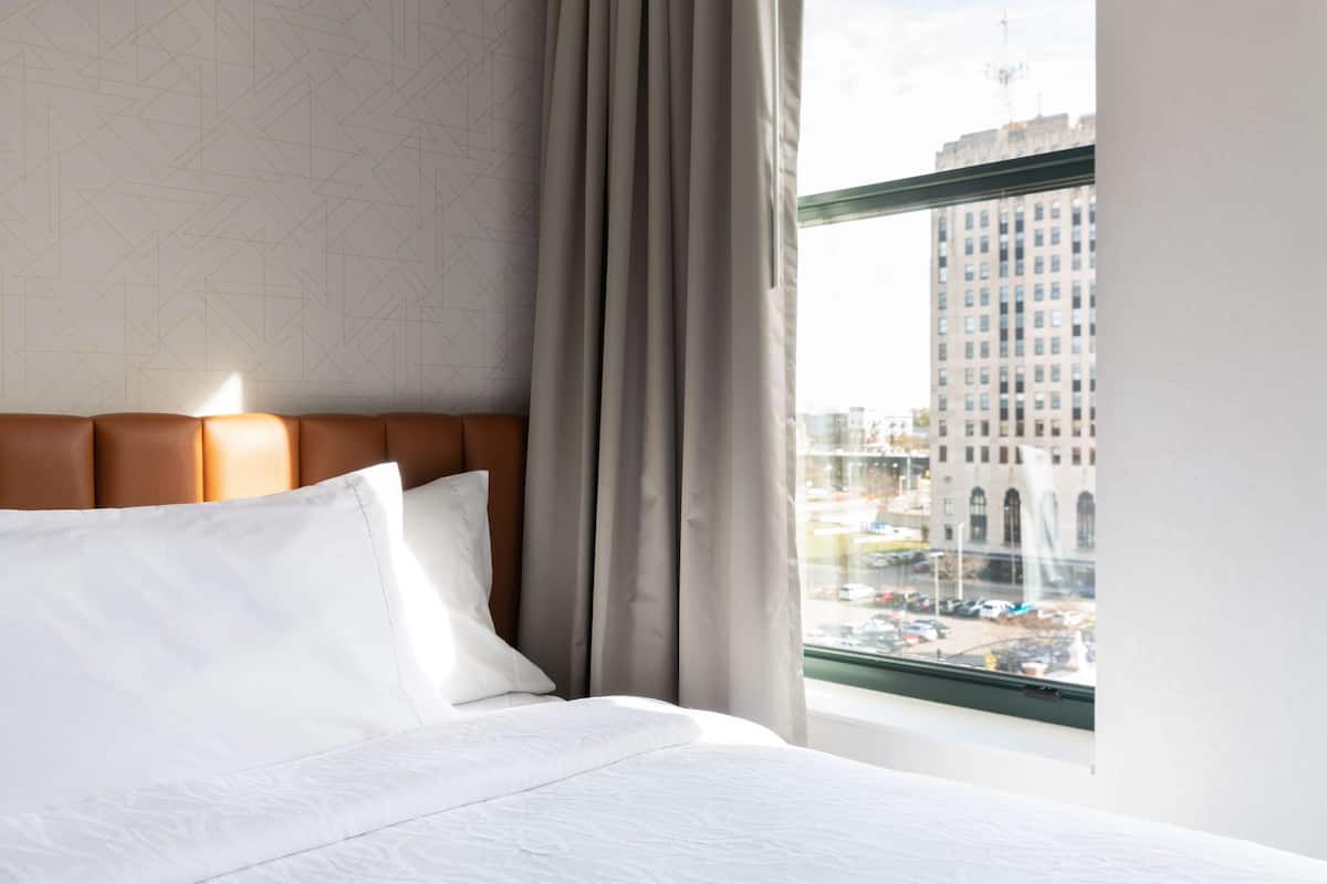 A hotel room with a large window, a bed with white linens, a headboard, and a geometric patterned wall.