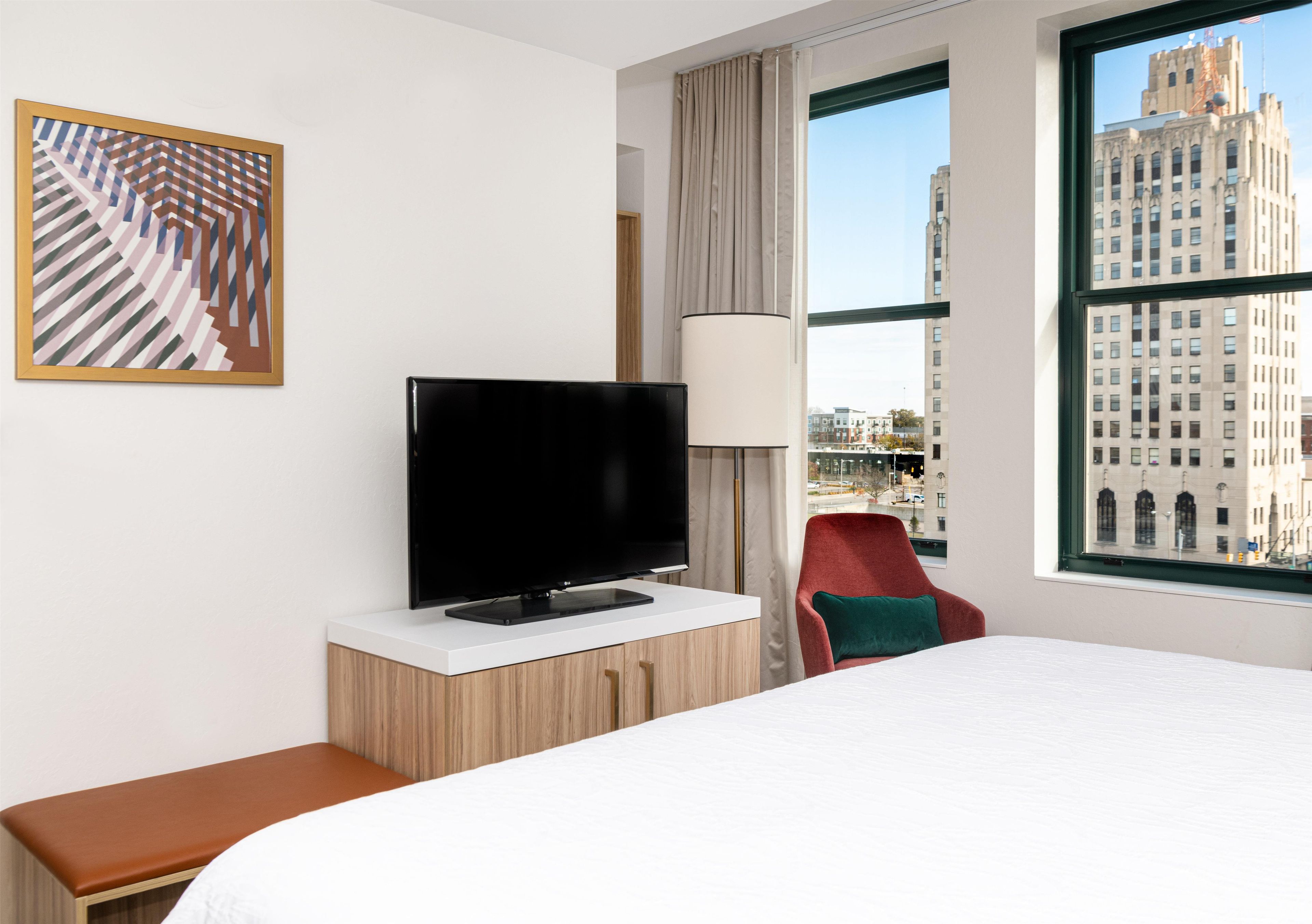 A hotel room with a bed, a television on a wooden cabinet, a red chair, a large window with a city view, and a framed abstract painting on the wall.