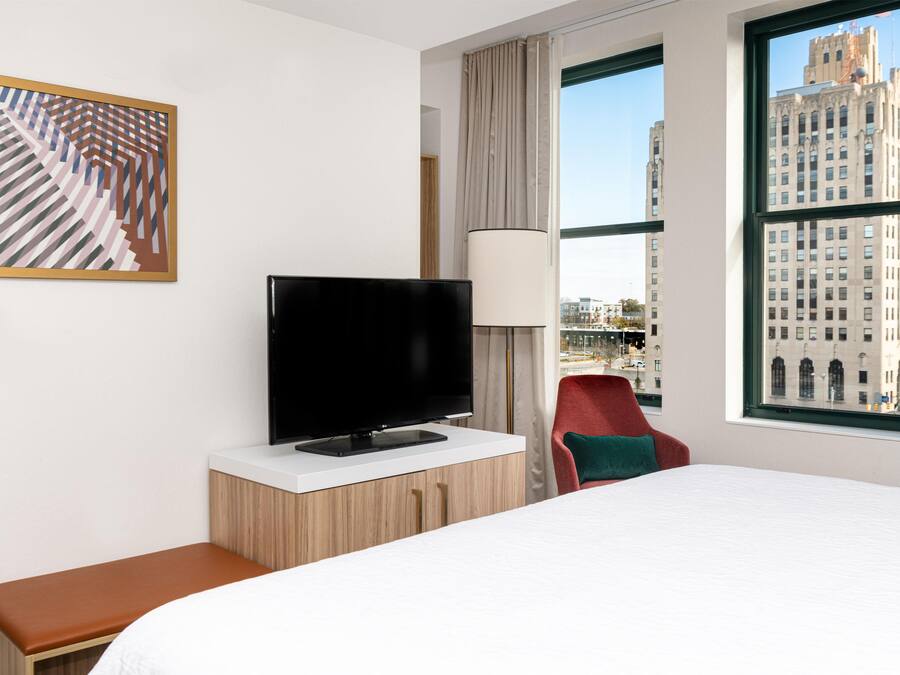 A hotel room with a bed, a television on a wooden cabinet, a red chair, a large window with a city view, and a framed abstract painting on the wall.