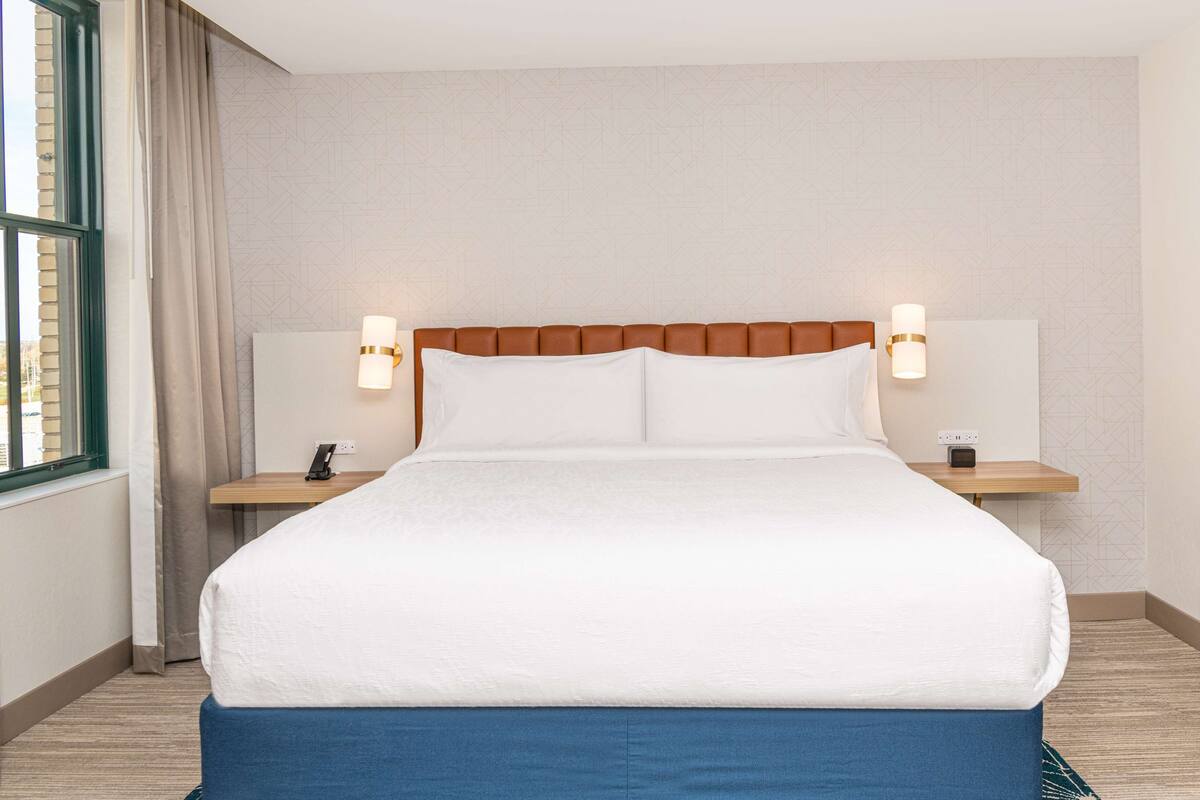 Una cama bien tendida con sábanas blancas, dos mesitas de noche con lámparas y vistas al exterior a través de una ventana con cortinas.