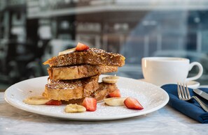 A stack of French toast with strawberries and bananas on a plate, a cup of coffee, and a fork on a napkin. - Hilton Garden Inn Flint Downtown (Flint)
