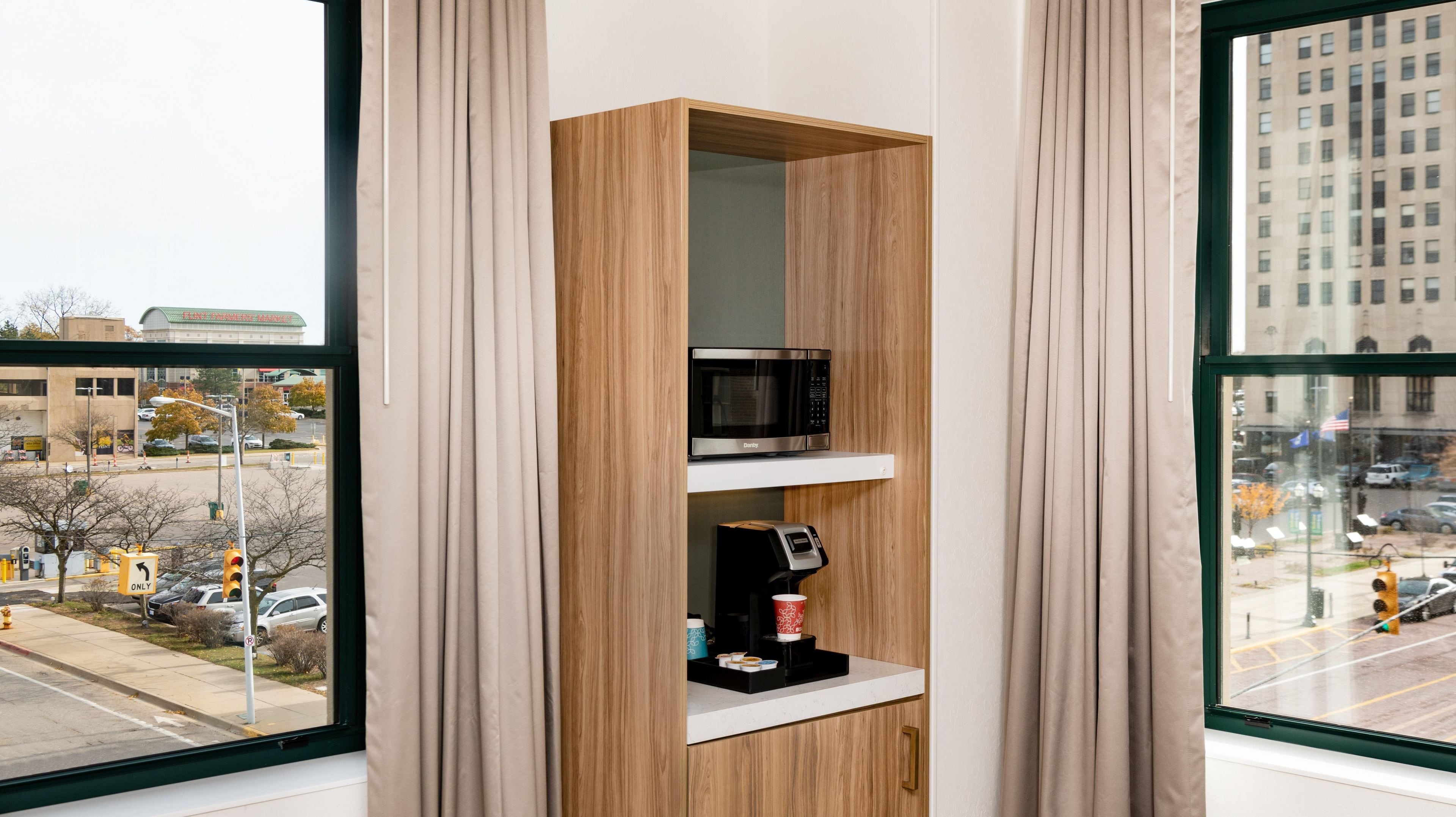 Una habitación con una estantería de madera que contiene un microondas y una cafetera, una ventana con vistas a un paisaje urbano y una puerta con el cartel de estacionamiento 'P'.