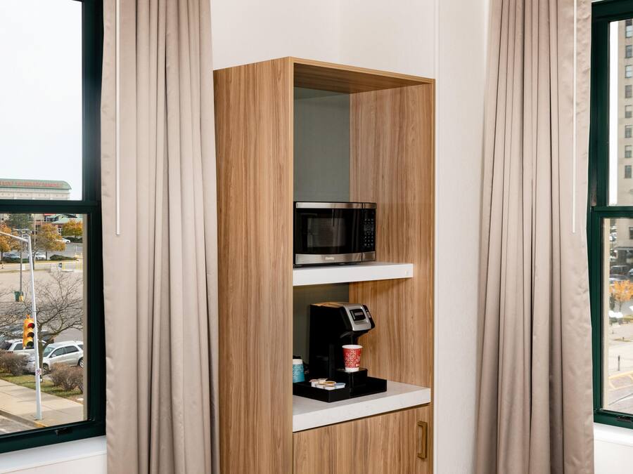 Una habitación con una estantería de madera que contiene un microondas y una cafetera, una ventana con vistas a un paisaje urbano y una puerta con el cartel de estacionamiento 'P'.