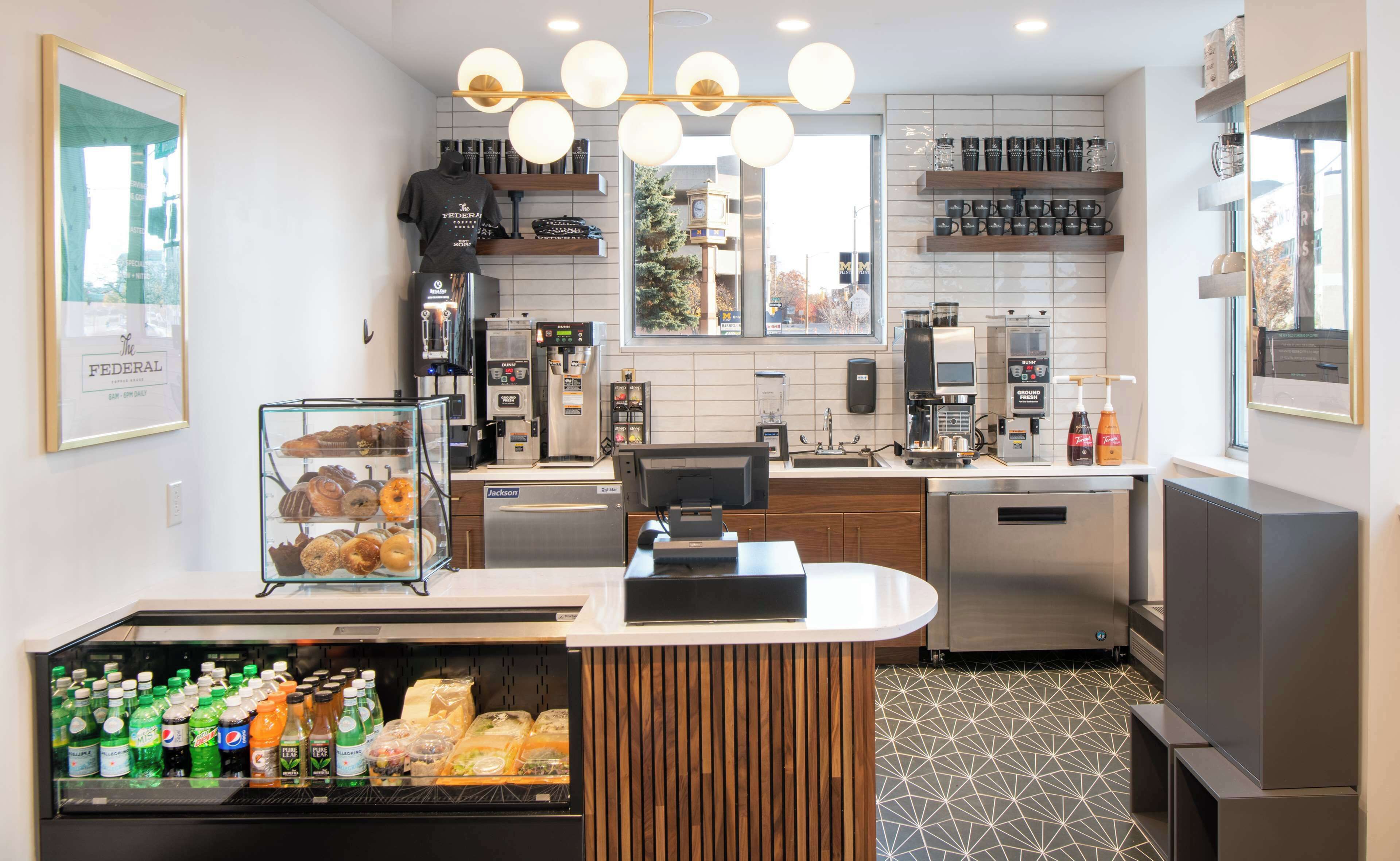 A modern coffee shop with a counter, coffee machines, a display of pastries, and a refrigerated beverage case.