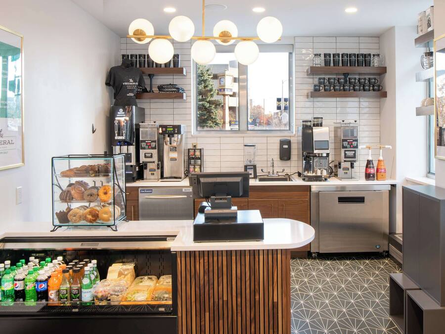 A modern coffee shop with a counter, coffee machines, a display of pastries, and a refrigerated beverage case.