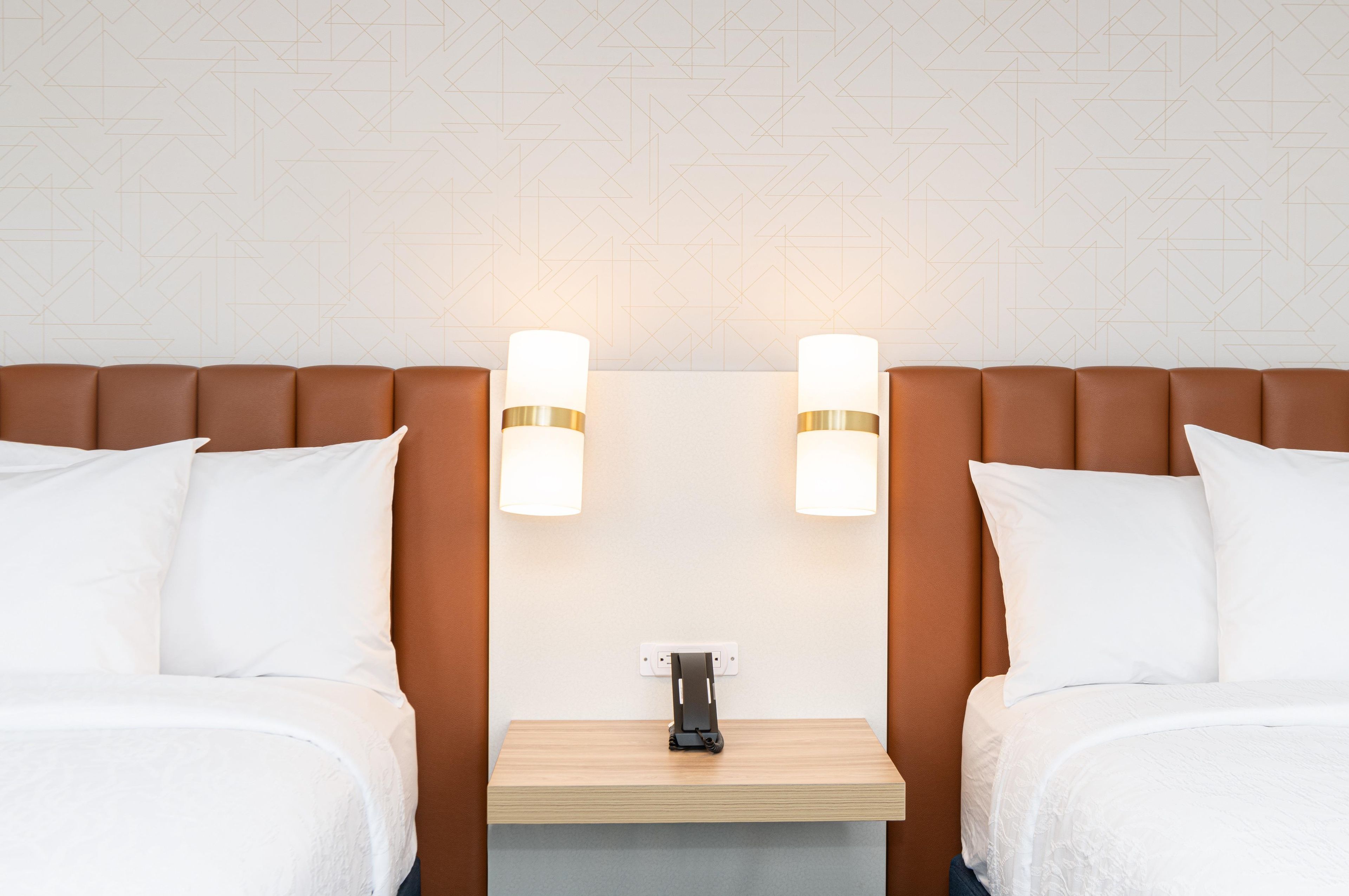 A hotel room with two beds, a wooden nightstand, and wall-mounted lamps.