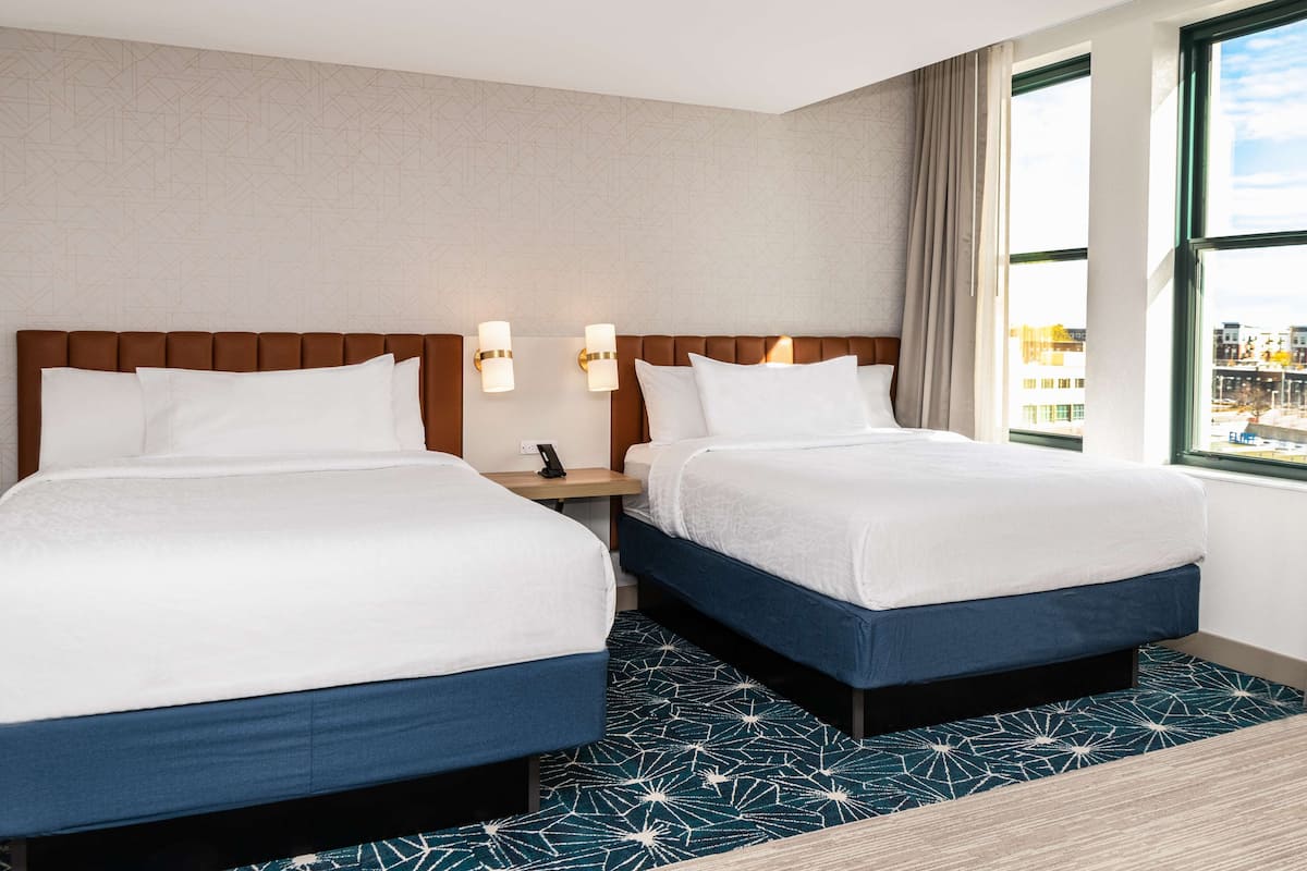 Habitación de hotel con dos camas, una mesita de noche, un teléfono y una ventana con vista a la ciudad.