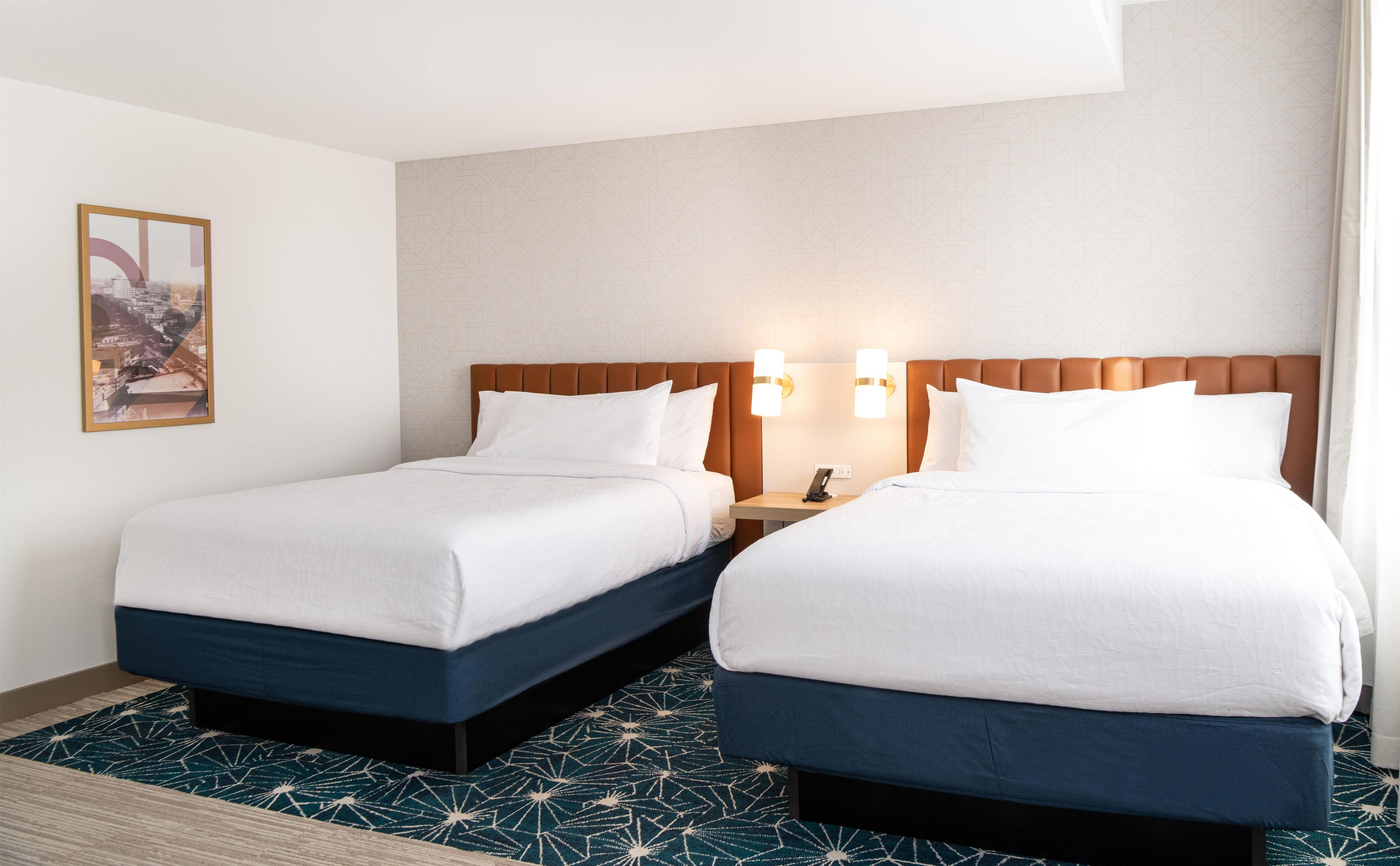 A hotel room with two beds, a nightstand, and a wall-mounted light fixture.