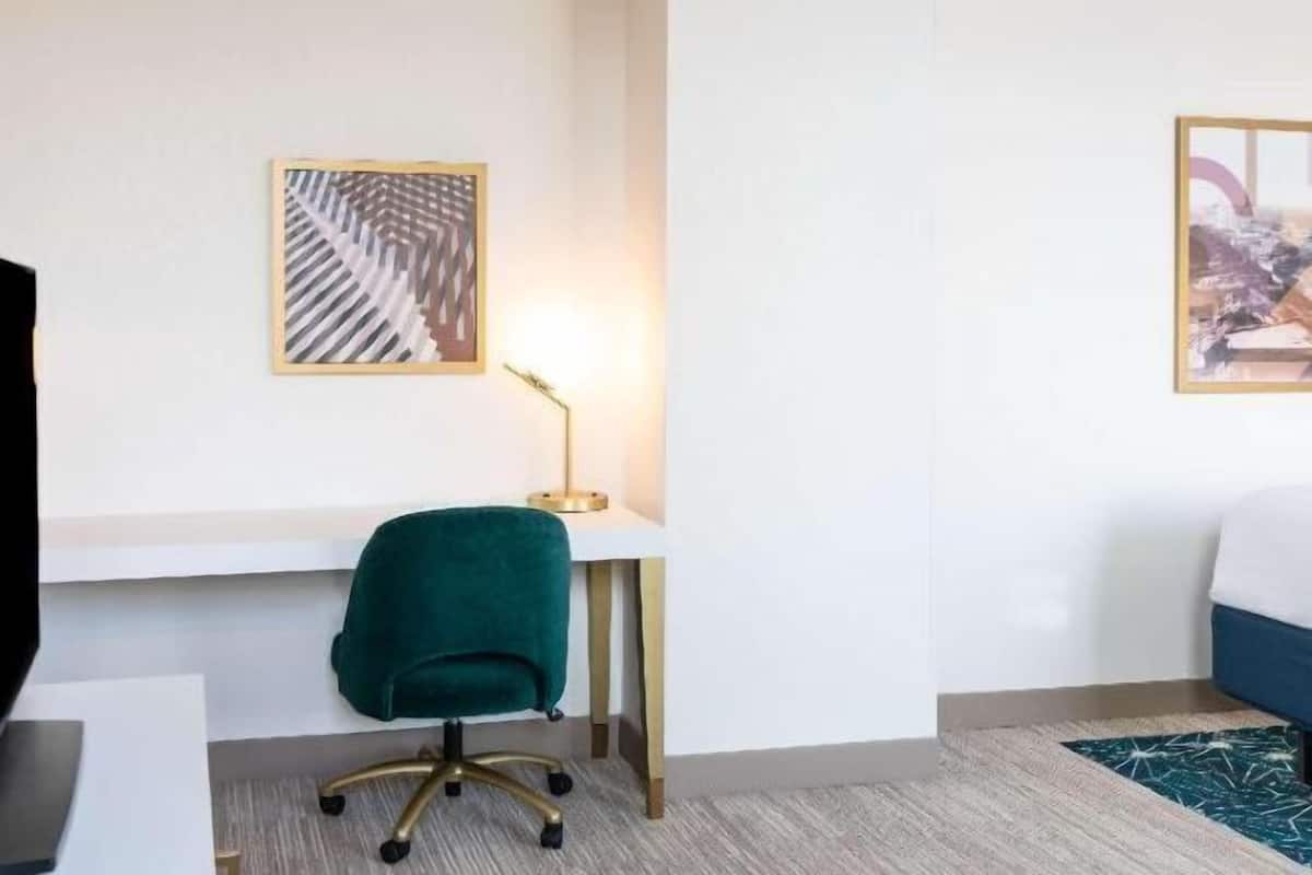 Una habitación de hotel con un escritorio, una silla verde, una lámpara y dos obras de arte en las paredes.