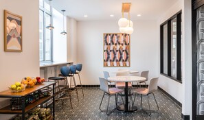 Un intérieur moderne avec un coin repas comprenant une table ronde et des chaises, un panier de fruits posé sur un comptoir, et un sol orné d’un motif géométrique.