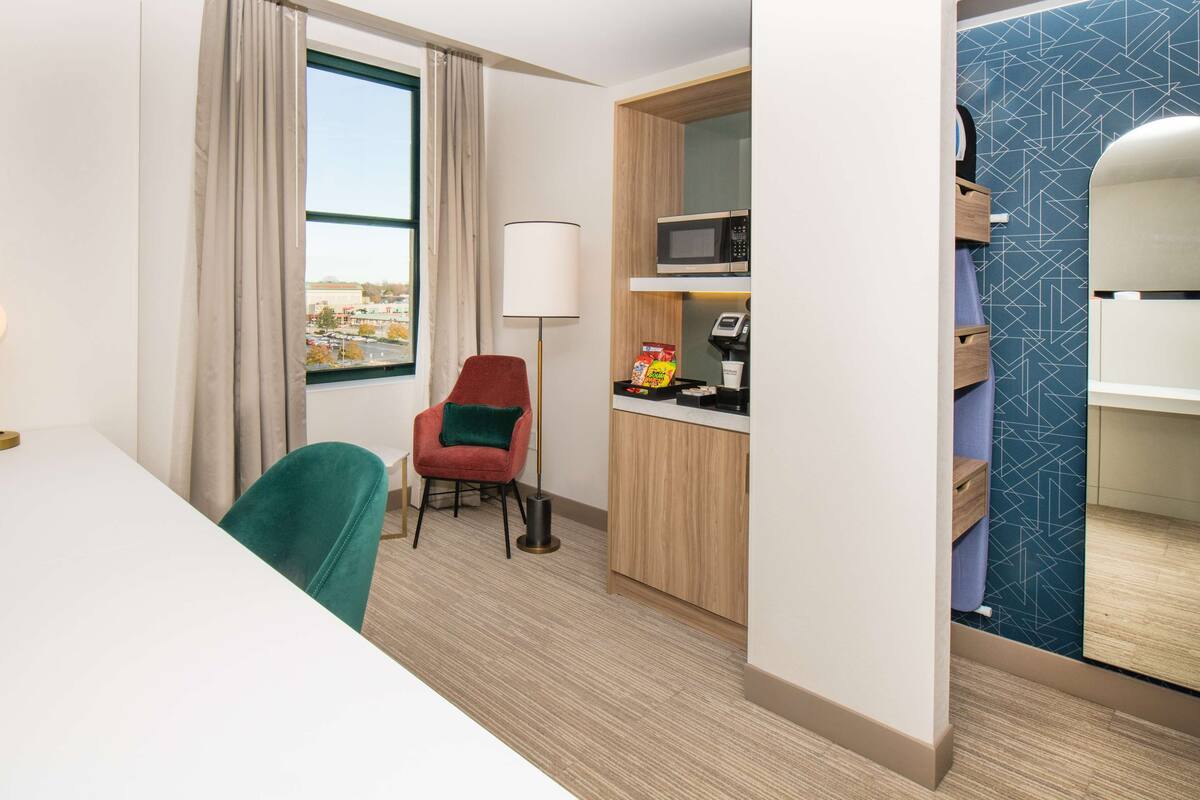 Habitación de hotel con escritorio, silla, lámpara, microondas, kitchenette pequeña y vistas a la ciudad.