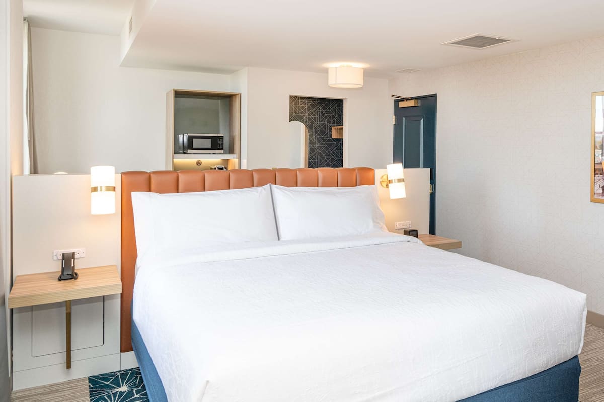 Habitación de hotel con cama, mesitas de noche, microondas, televisor montado en la pared y un cuadro enmarcado.