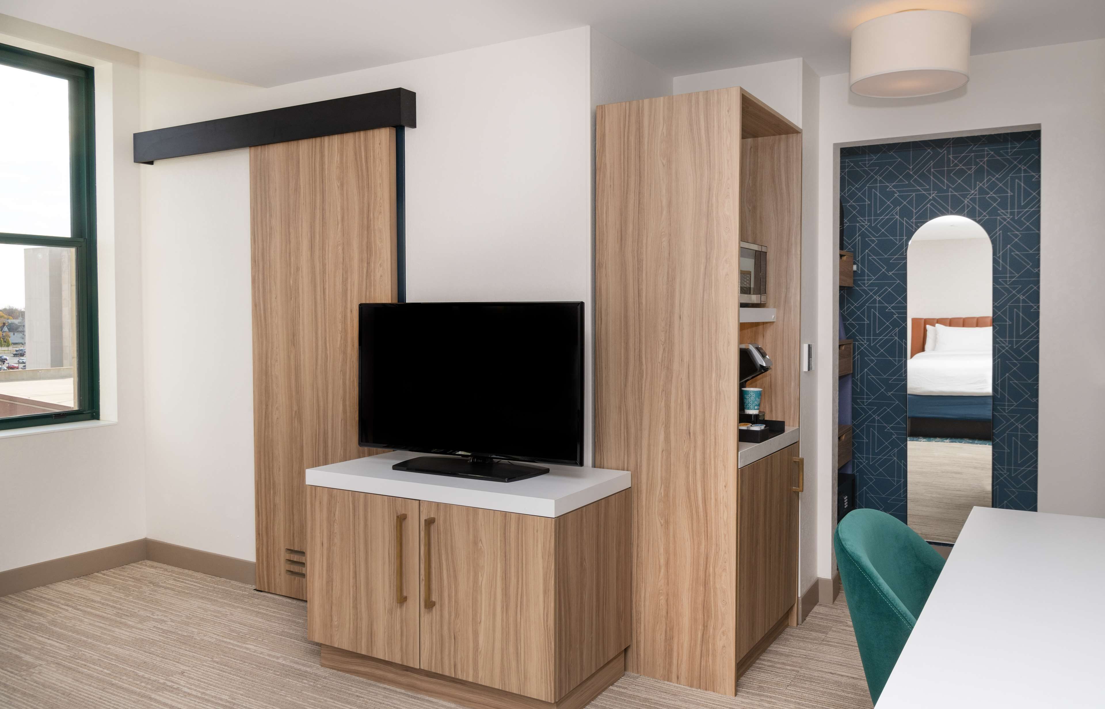 Una habitación moderna con una televisión sobre un mueble de madera, una mesa blanca y una silla color turquesa.
