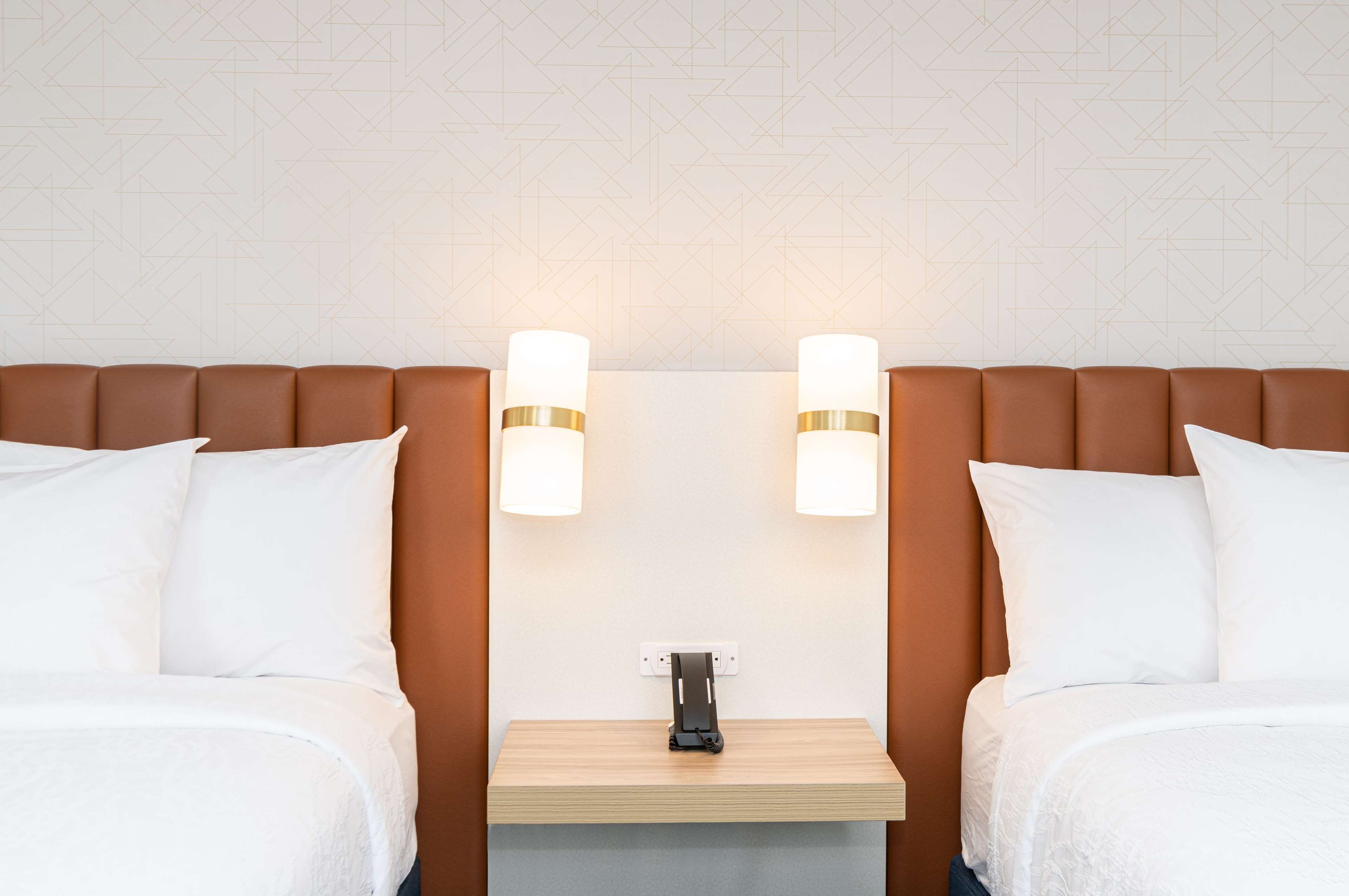 Quarto de hotel com duas camas, uma mesa de cabeceira de madeira e luminárias fixadas na parede.