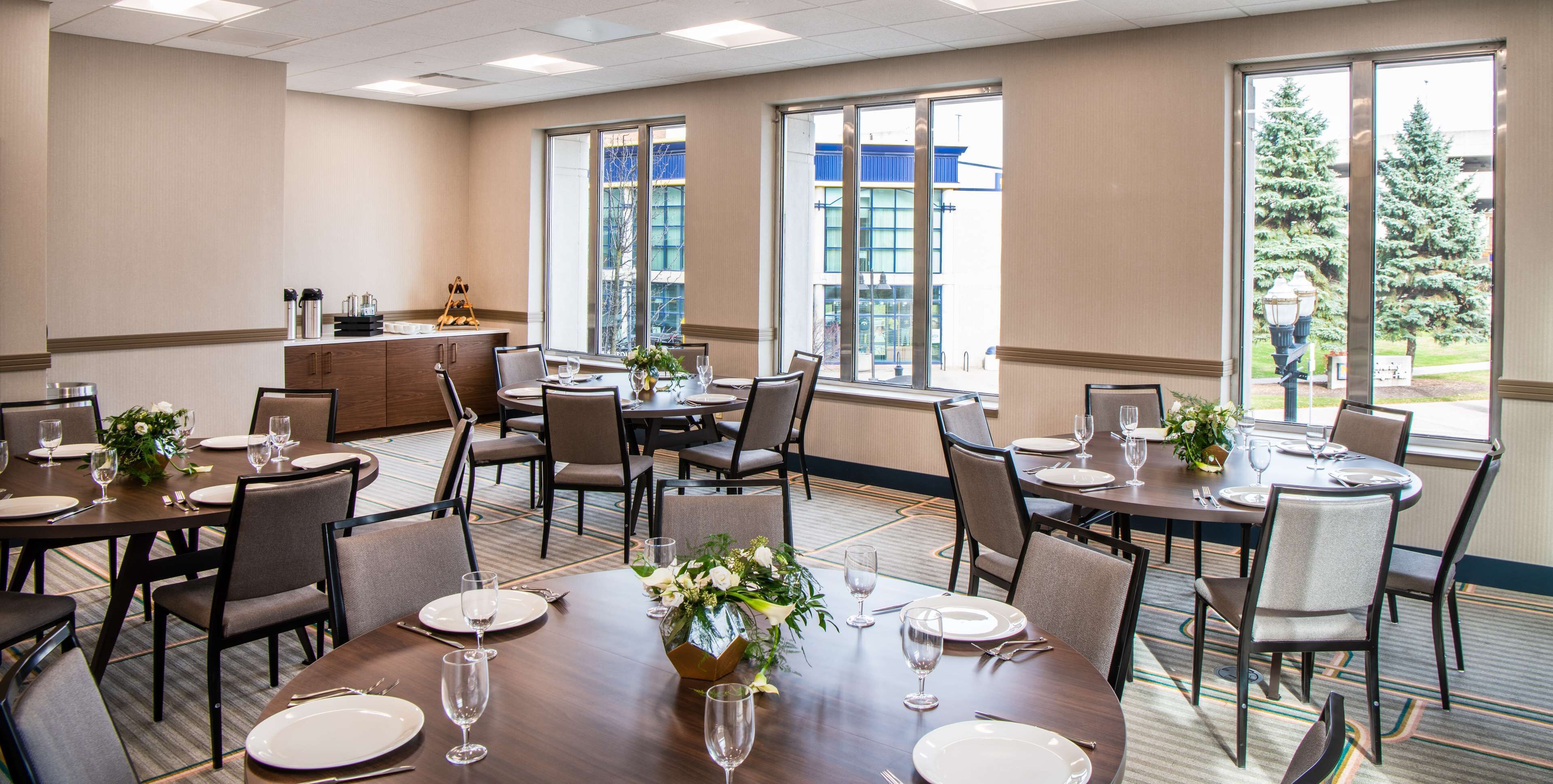 A hotel dining area with multiple tables set for dining, large windows providing natural light, and a view of the outside.
