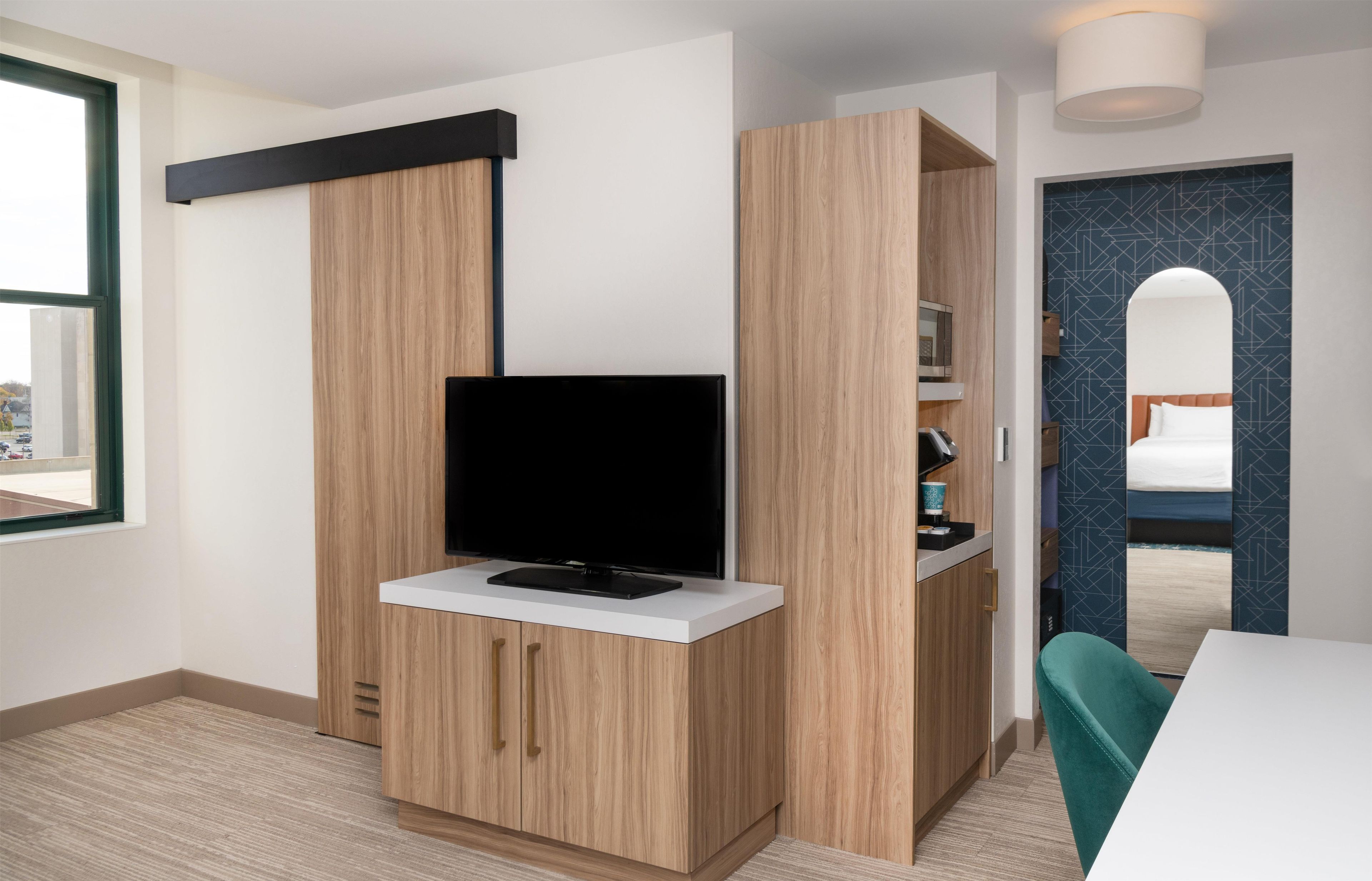 Habitación de hotel moderna con televisor de pantalla plana sobre un mueble blanco, armario de madera y vistas a la calle a través de un ventanal grande.
