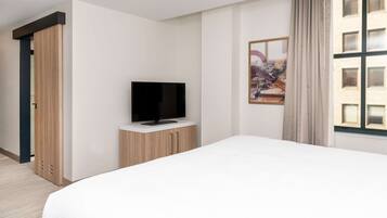 Habitación de hotel con cama, televisor en mueble de madera, ventana con cortinas e imagen de paisaje urbano en la pared.