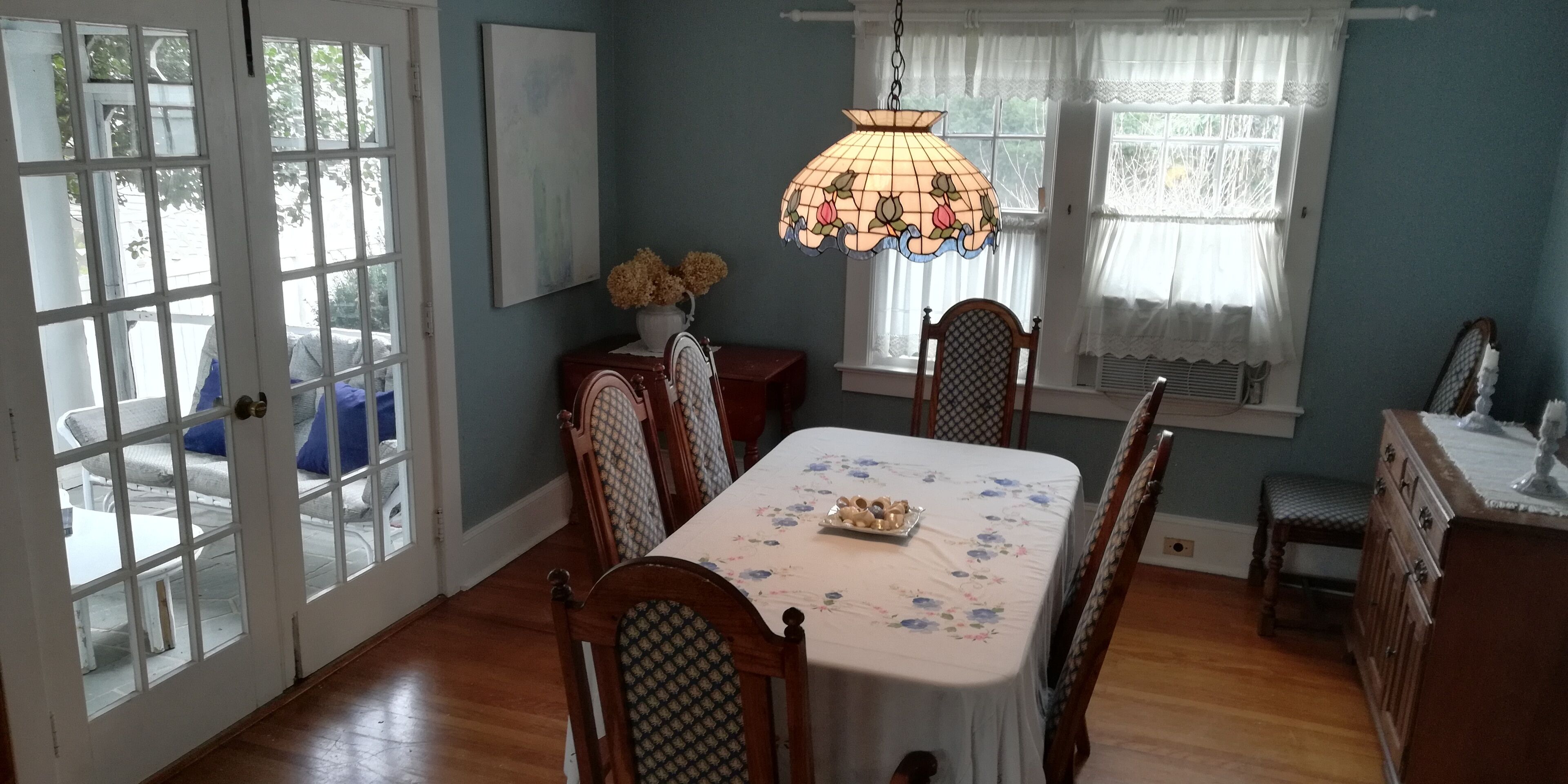 Une salle à manger avec une table dressée pour quatre personnes, un lustre et une fenêtre ornée de rideaux en dentelle.