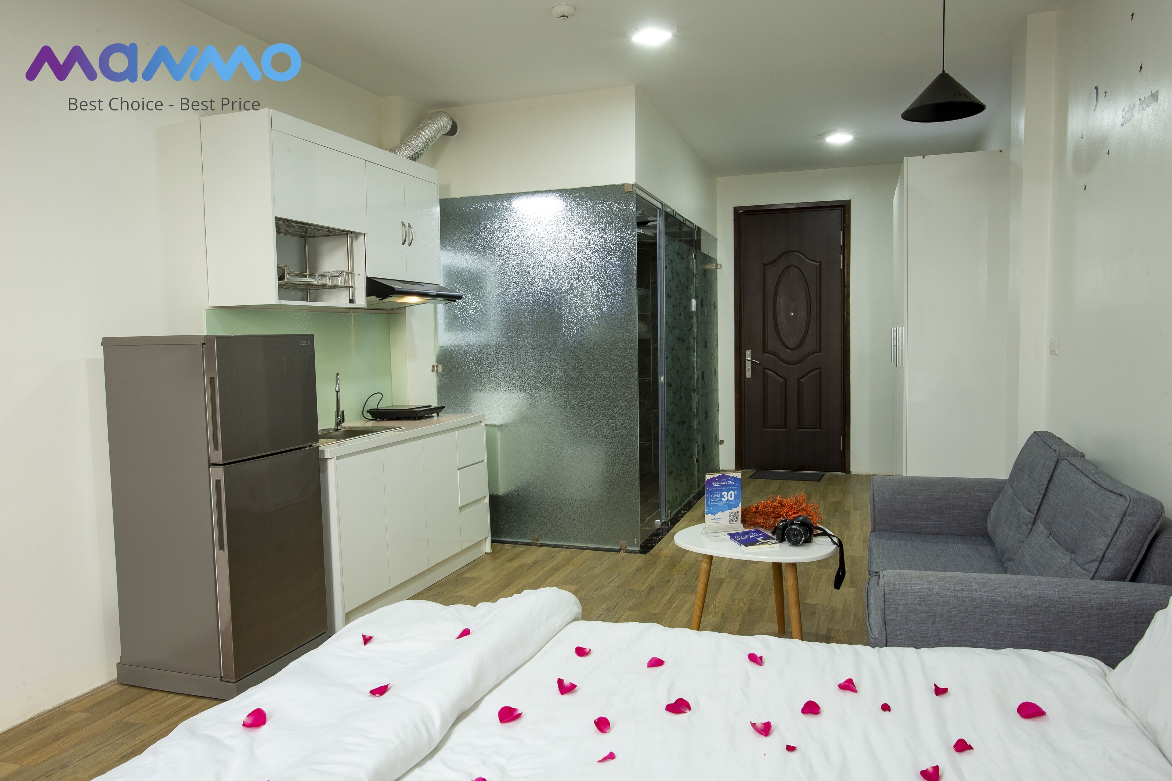 A compact hotel room with a kitchenette, a bed with rose petals, a grey sofa, and a small table with a sign and headphones.