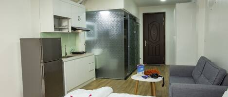 A compact hotel room with a kitchenette, a bed with rose petals, a grey sofa, and a small table with a sign and headphones.
