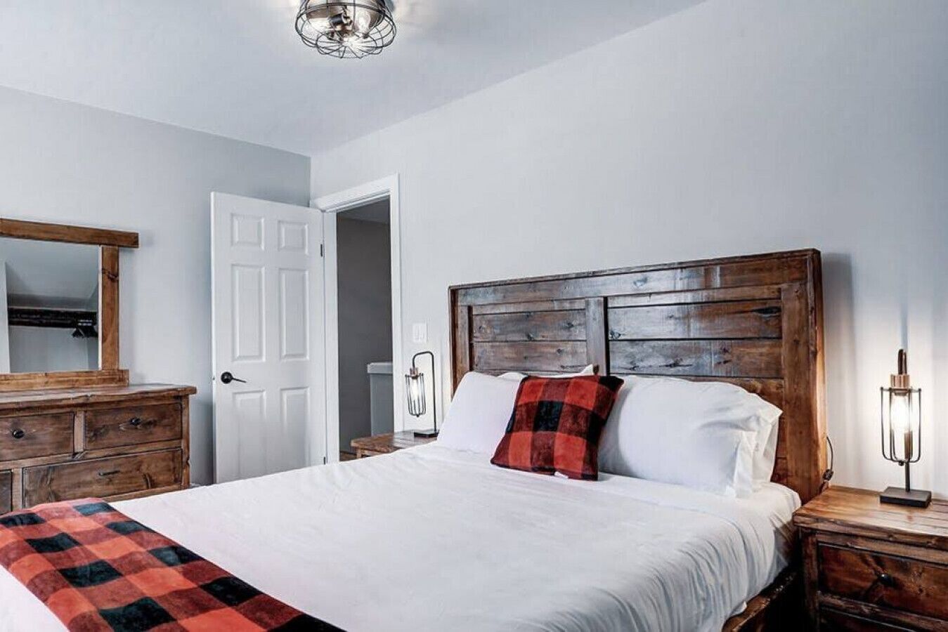 Quarto com cabeceira de madeira, colcha branca e manta xadrez vermelha e preta.