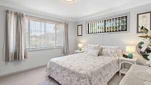 A bedroom with a large bed, two bedside tables with lamps, a window with curtains, and a view of greenery outside. - Duplex with views in a quiet area (Nelson Bay)