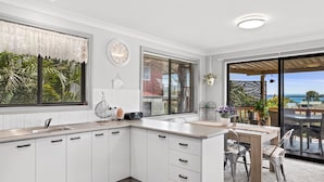 A modern kitchen with white cabinets, a large window with blinds, a wall clock, and a view of the ocean through a sliding glass door. - Duplex with views in a quiet area (Nelson Bay)