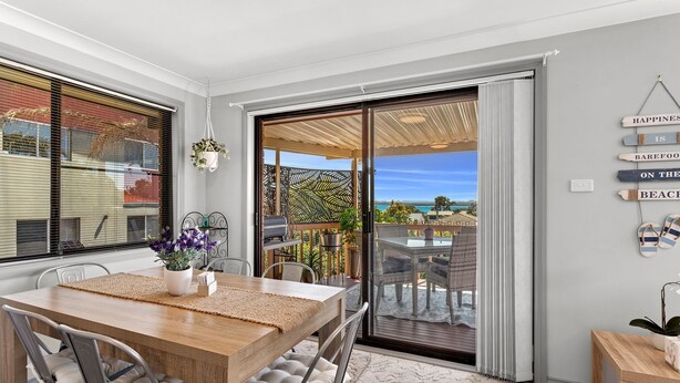 A dining area with a wooden table and chairs, a view of the beach, and a sign on the wall that reads "Happiness is barefoot on the beach." - Duplex with views in a quiet area (Nelson Bay)