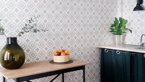 Eine Küche mit gemustertem Wandpapier, einem Holztisch mit Obstschale, einem grünen Blumenvasen, einer Hängelampe und einem grünen Vorhang.
