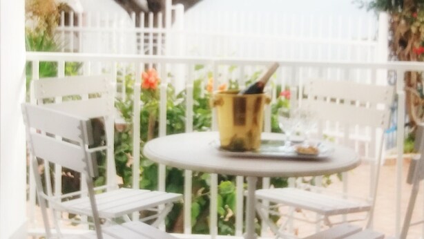 Ein Tisch und Stühle aus weißem Metall auf einem Balkon mit einem weißen Geländer, einem Blumentopf und einem Gebäude im Hintergrund.