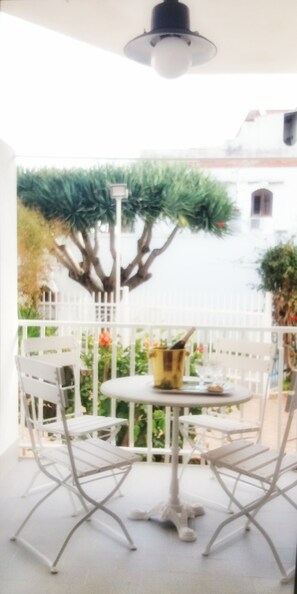 Ein Tisch und Stühle aus weißem Metall auf einem Balkon mit einem weißen Geländer, einem Blumentopf und einem Gebäude im Hintergrund.