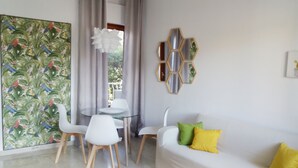 Ein modernes Wohnzimmer mit einem Glastisch, weißen Stühlen, einem weißen Sofa mit gelben Kissen, einem großen tropischen Wandbild und einem sechseckigen Spiegel an der Wand.