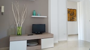 Ein modernes Wohnzimmer mit einem Flachbildfernseher auf einer Holztruhe, weißen Regalen mit Dekoration und einem Gemälde an der Wand.