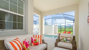 Un espace patio ensoleillé avec un canapé en osier, des coussins colorés, un fauteuil en osier et une vue sur une piscine et la verdure environnante.