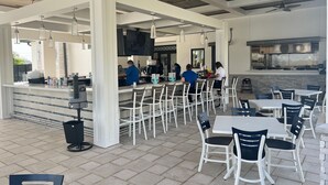 Un café en plein air avec des tables et des chaises blanches, un coin bar et une cuisine à l’arrière-plan.