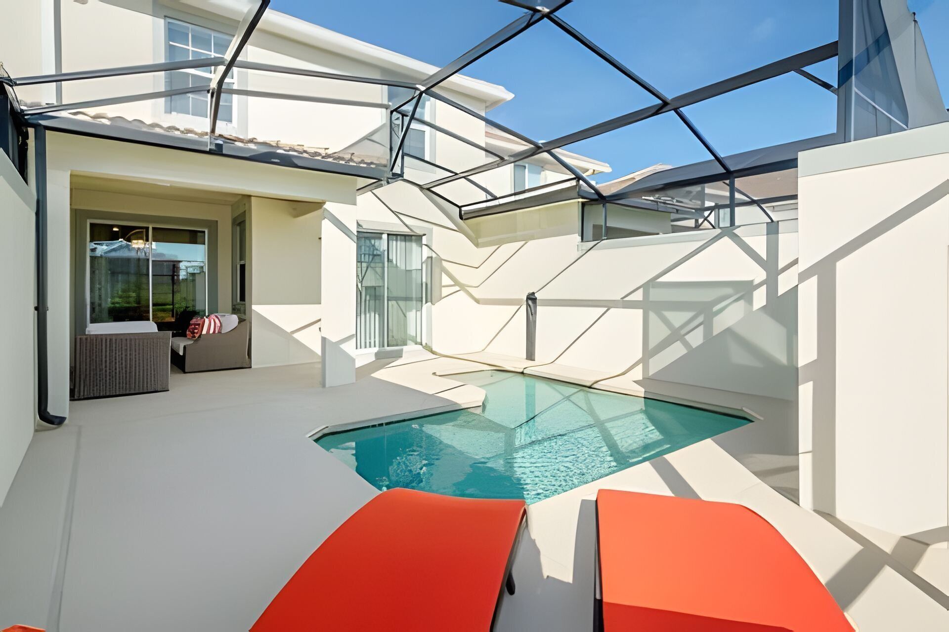 A modern outdoor pool area with glass walls, a seating area with orange loungers, and a clear view of the surrounding landscape.