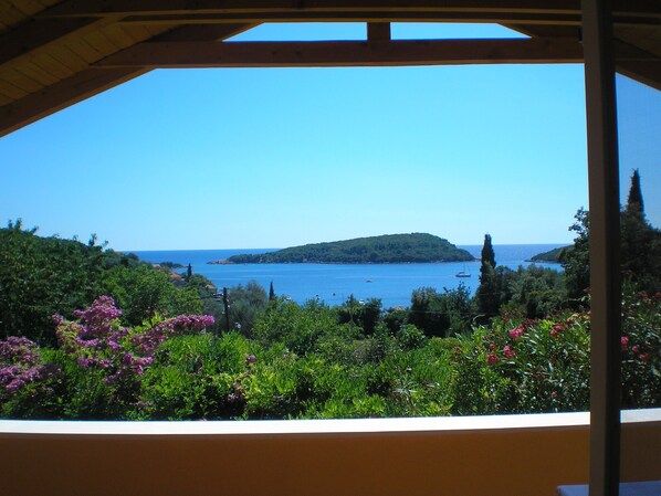 A view from a covered area overlooking a coastal landscape with a small island, lush greenery, and a clear blue sky.