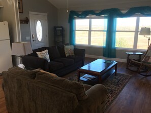 A living room with a dark brown sofa, a coffee table, and a rocking chair. There is a window with blue curtains, a door with a glass panel, and a small table with a lamp. - Our Cozy, Quiet, Comfortable place in the Country - 2 Bedroom 900 sq ft w/view (Lockhart)