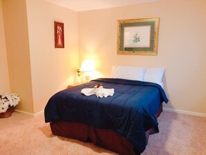 A neatly made bed with a blue cover, white pillows, and a small bouquet of flowers on the bed. There is a lamp on a bedside table and a framed picture on the wall. - Beautiful apartment in Historic downtown location close to everything ! (Greensboro)