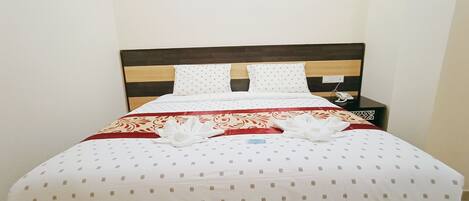A neatly made bed with a patterned bedspread, two pillows, and a red and white decorative pillow. There is a small bedside table with a phone and a wall-mounted light switch.