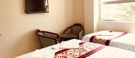 A hotel room with two beds, a TV mounted on the wall, and a window with a view of greenery.