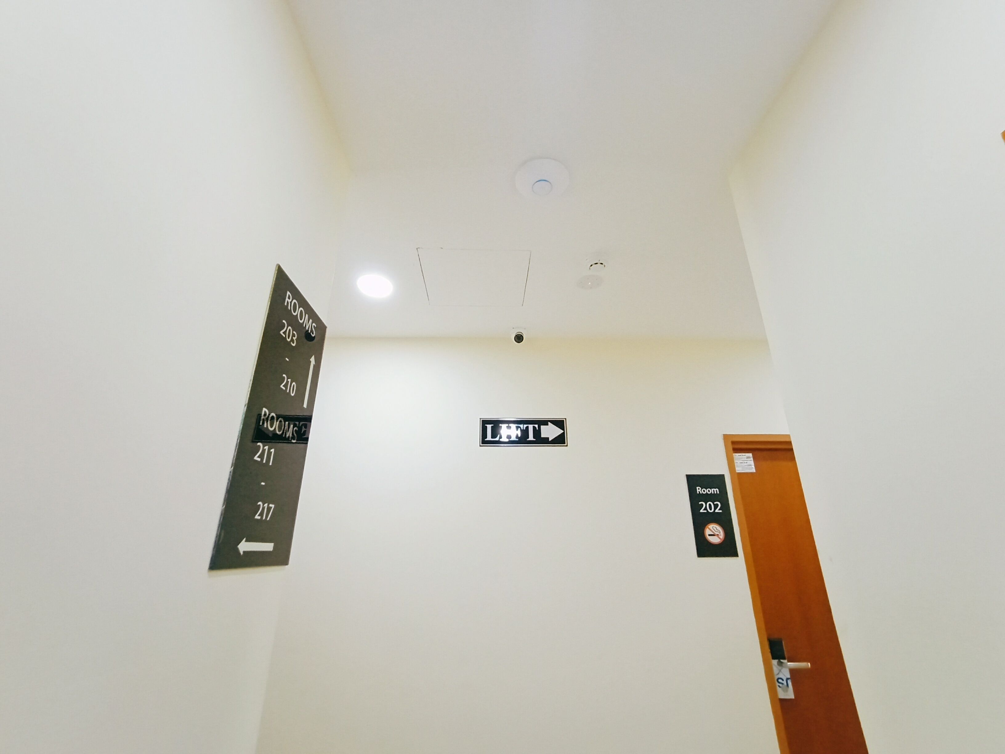 Corredor de hotel com placas de direção para os quartos 203, 210, 211 e 217, e uma placa indicando a localização do elevador.
