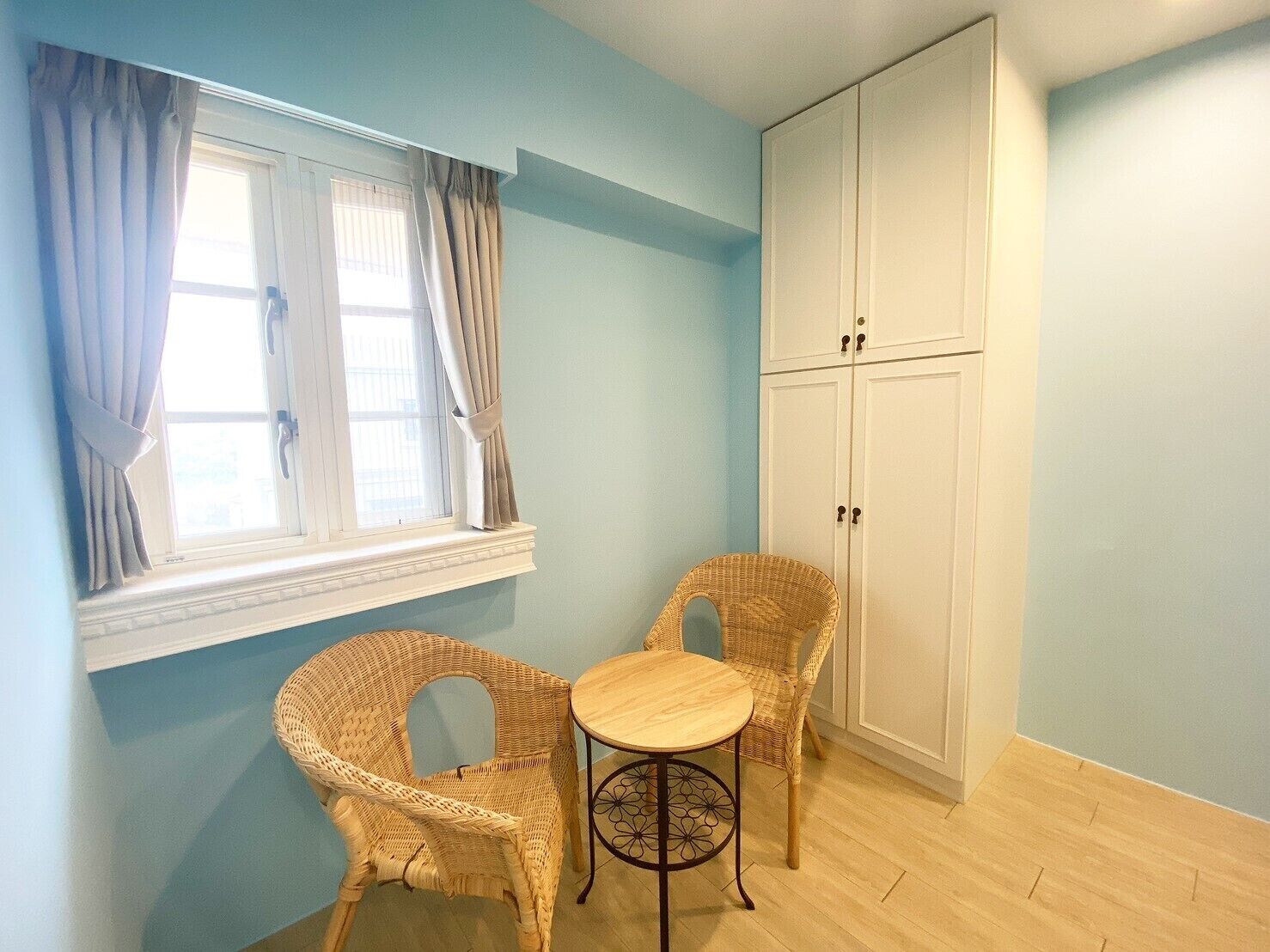 A room with light blue walls, a white wardrobe, two wicker chairs, a small round table, and a window with curtains.