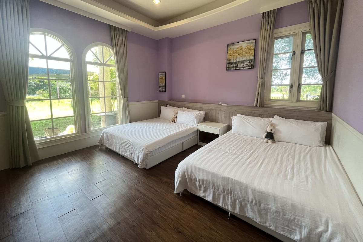 A room with two single beds, a nightstand, a painting, and large windows with curtains.