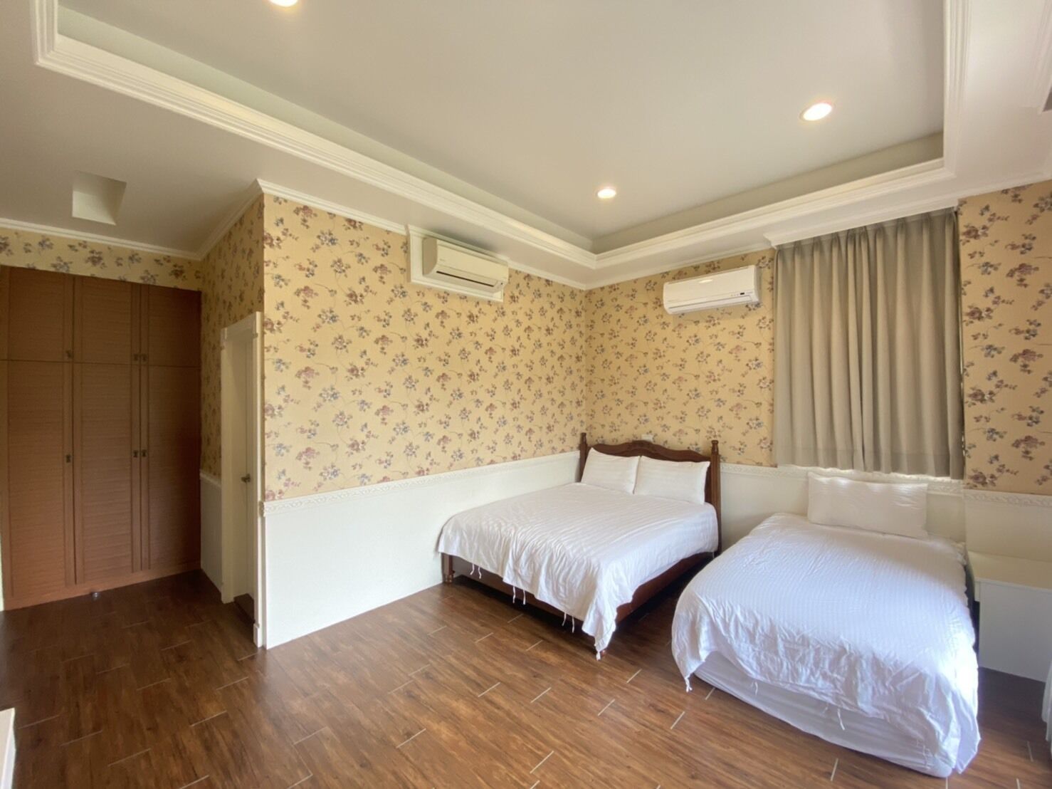 Un dormitorio con dos camas, suelo de madera, papel tapiz floral y un equipo de aire acondicionado.