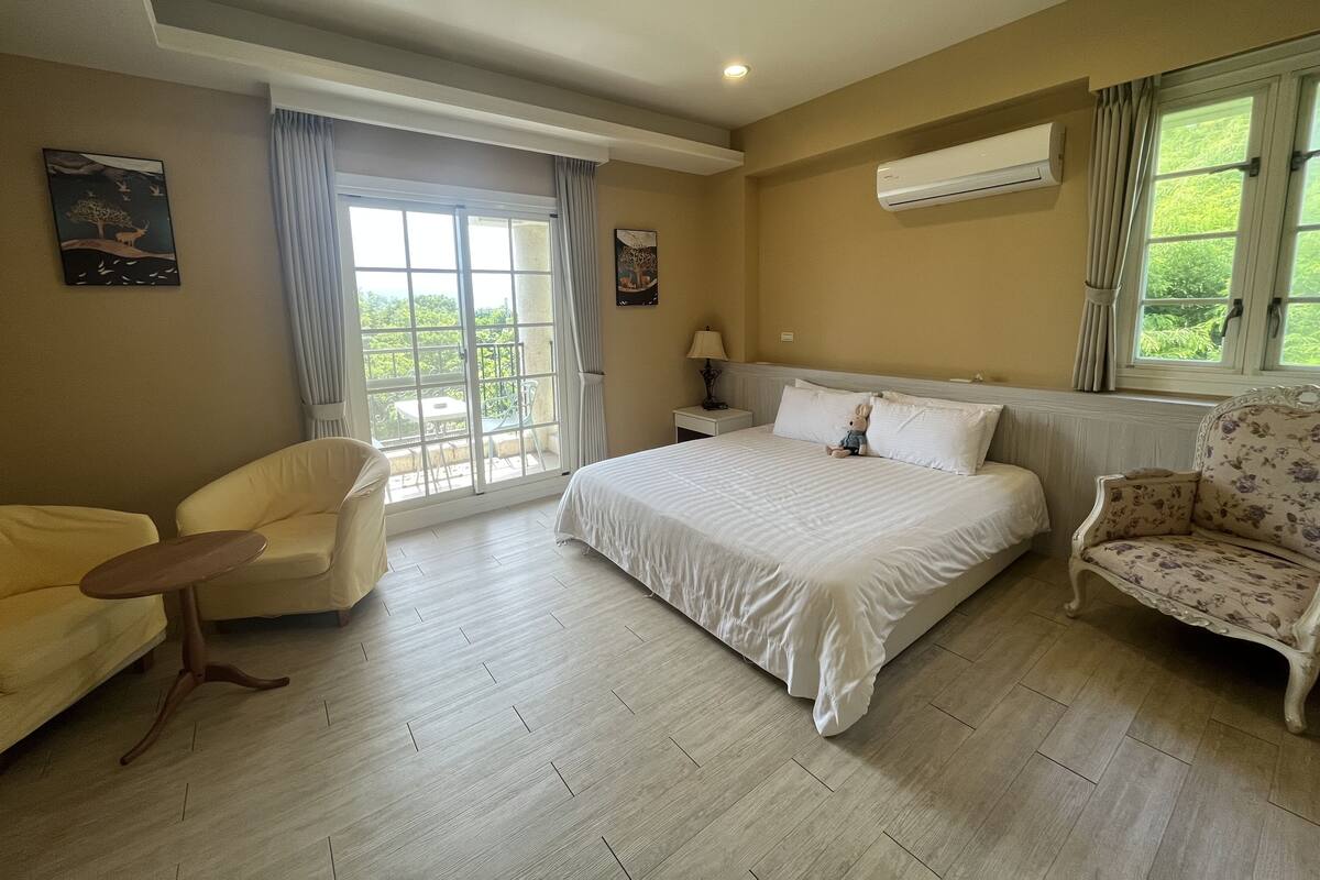 A bedroom with a large bed, two chairs, a small table, and a window with curtains.