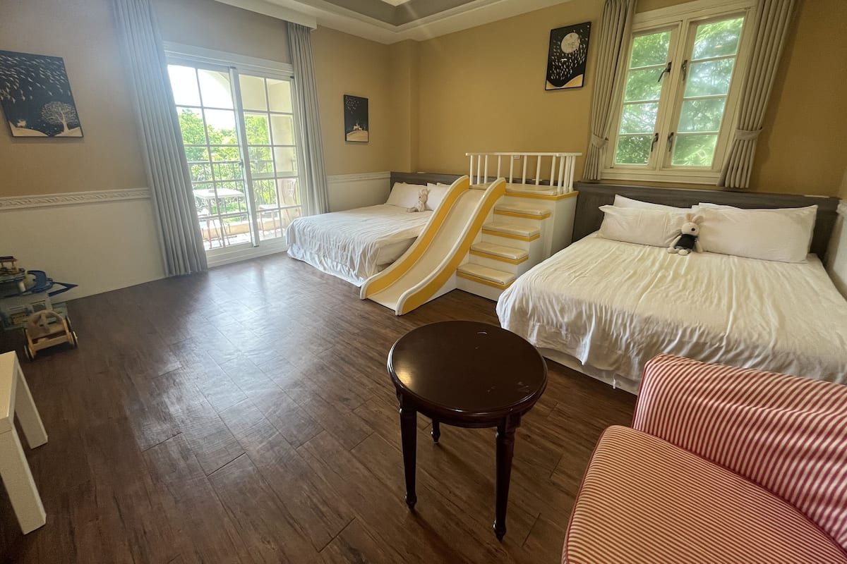 A hotel room with two beds, a small table, a sofa, a window with curtains, and a view of greenery outside.