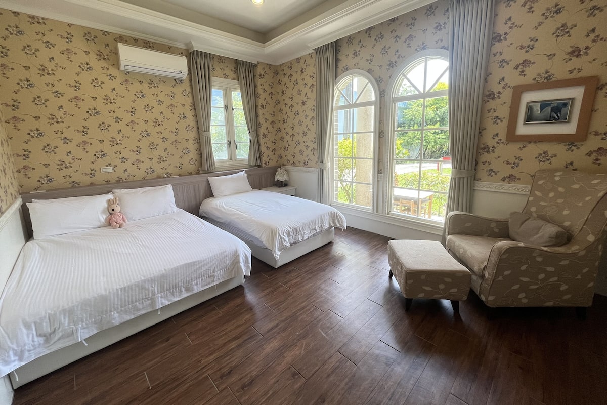 A hotel room with two beds, a chair, a window with curtains, and a framed picture on the wall.